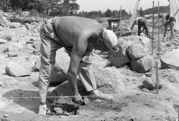 Svartvit bild på en arkeolog som står böjd och gräver med en murarslev. Stenblock och andra arkeologer i bakgrunden.