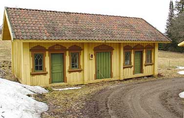 Gul träbyggnad med gröna dörrar och bruna foder runt fönster och dörrar.